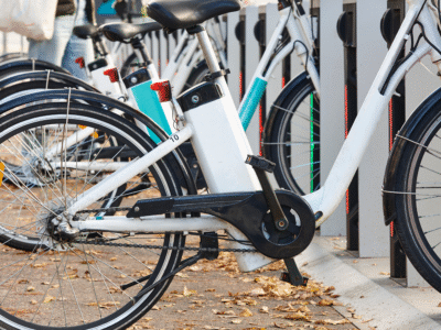 A row of white electric bicycles parked and locked in a public bike share docking station on a city sidewalk with fallen leaves on the ground. The image relates to California’s new e-bike classification laws and how updated micromobility regulations may affect electric bicycle riders.