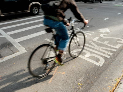 A cyclist rides through an intersection with marked bike lanes and crosswalk lines. The photo represents California’s new daylighting law, which increases visibility near crosswalks to enhance pedestrian and cyclist safety.