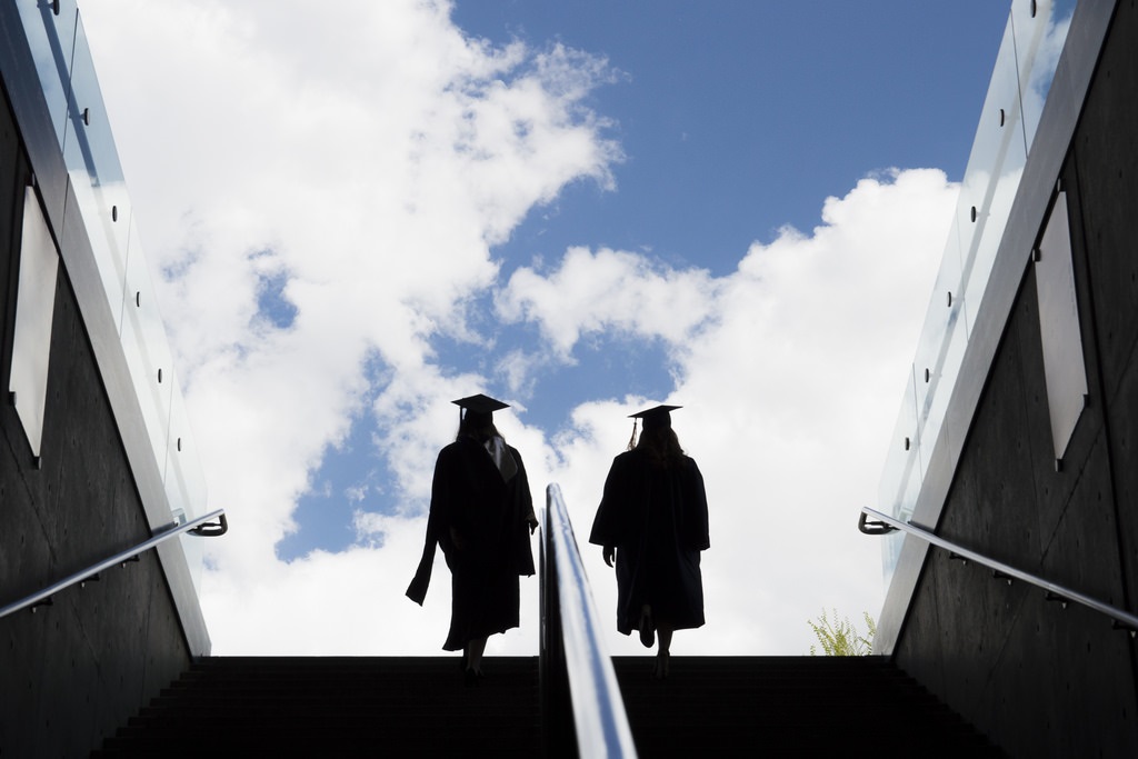Two graduates in a cap and gown at the top of a tall stair case with beautiful blue skies behind them.