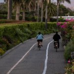 Two people ride bicycles along a paved coastal path lined with palm trees, colorful flowers, and lush greenery at sunset. To the right, a beach and ocean stretch into the distance beneath a soft pastel sky, with mountains visible on the horizon