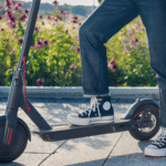 erson wearing black high-top sneakers and rolled-up jeans standing on a black electric scooter on a paved path, with blooming purple flowers and greenery in the background