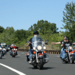 A group of motorcyclists ride together on a multi-lane road during daytime, with riders wearing helmets and casual clothing. Trees line the background, and a few cars follow behind in the right lane under a clear blue sky