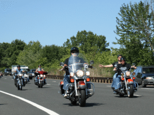 A group of motorcyclists ride together on a multi-lane road during daytime, with riders wearing helmets and casual clothing. Trees line the background, and a few cars follow behind in the right lane under a clear blue sky